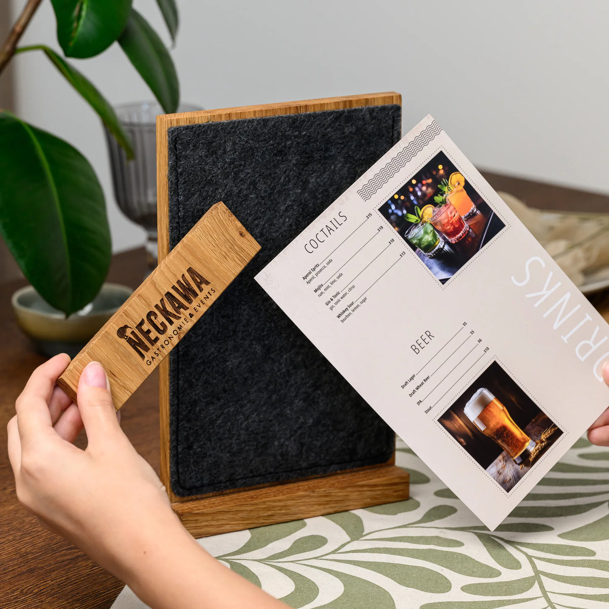 Hands holding open a table tent, revealing the engraved oak bar and felt panel with a menu page inside 