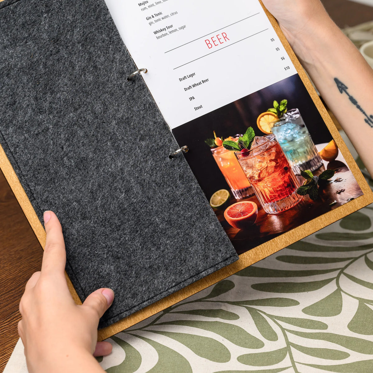 Hands holding open a ring-bound menu board, revealing a printed menu page with colorful beverage photos 