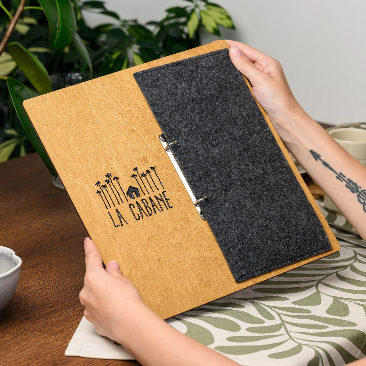 Hands holding open a menu board, showing the engraved plywood front and felt cover panel near a plant 