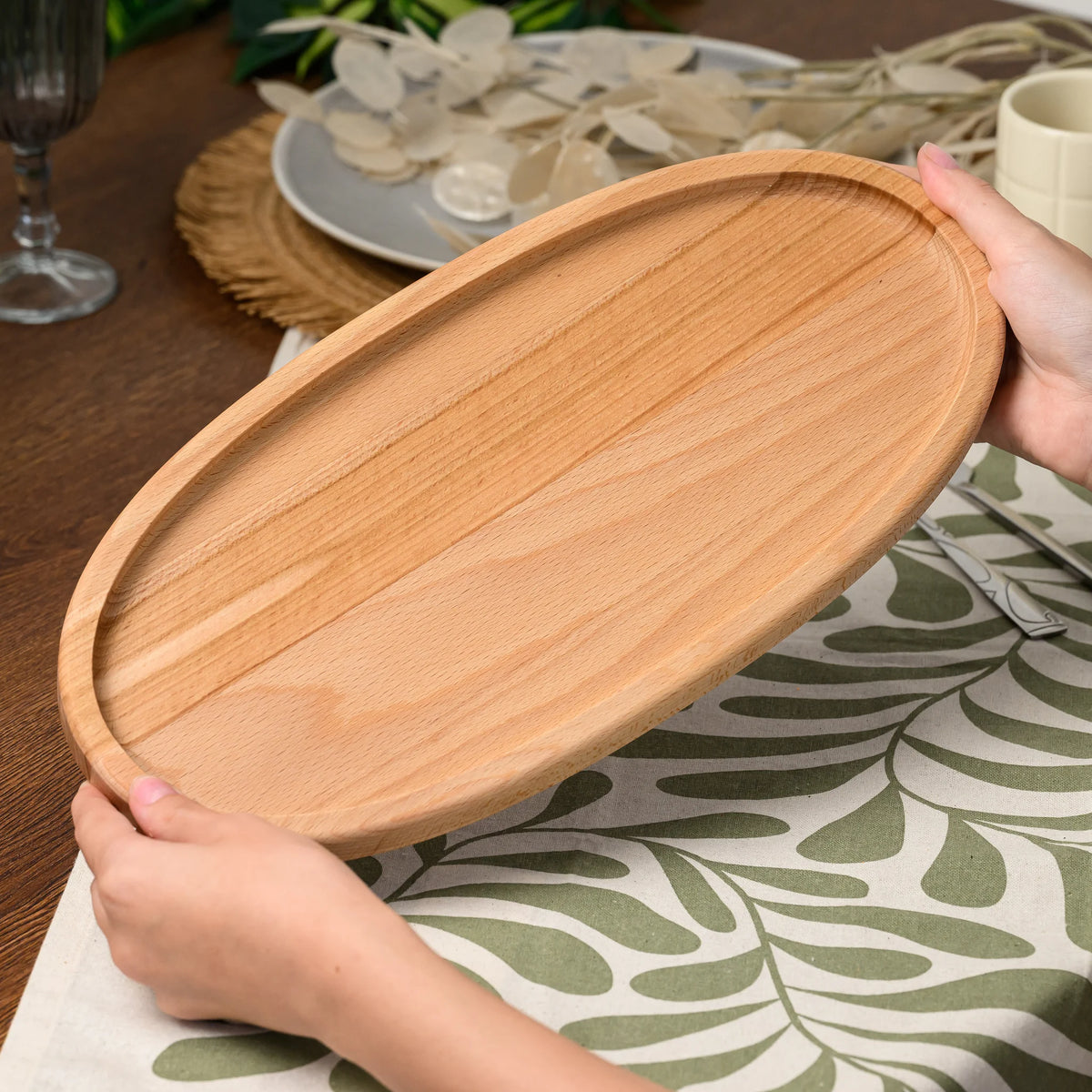 Hands holding the beech wood oval serving board at an angle above a table set with a wine glass