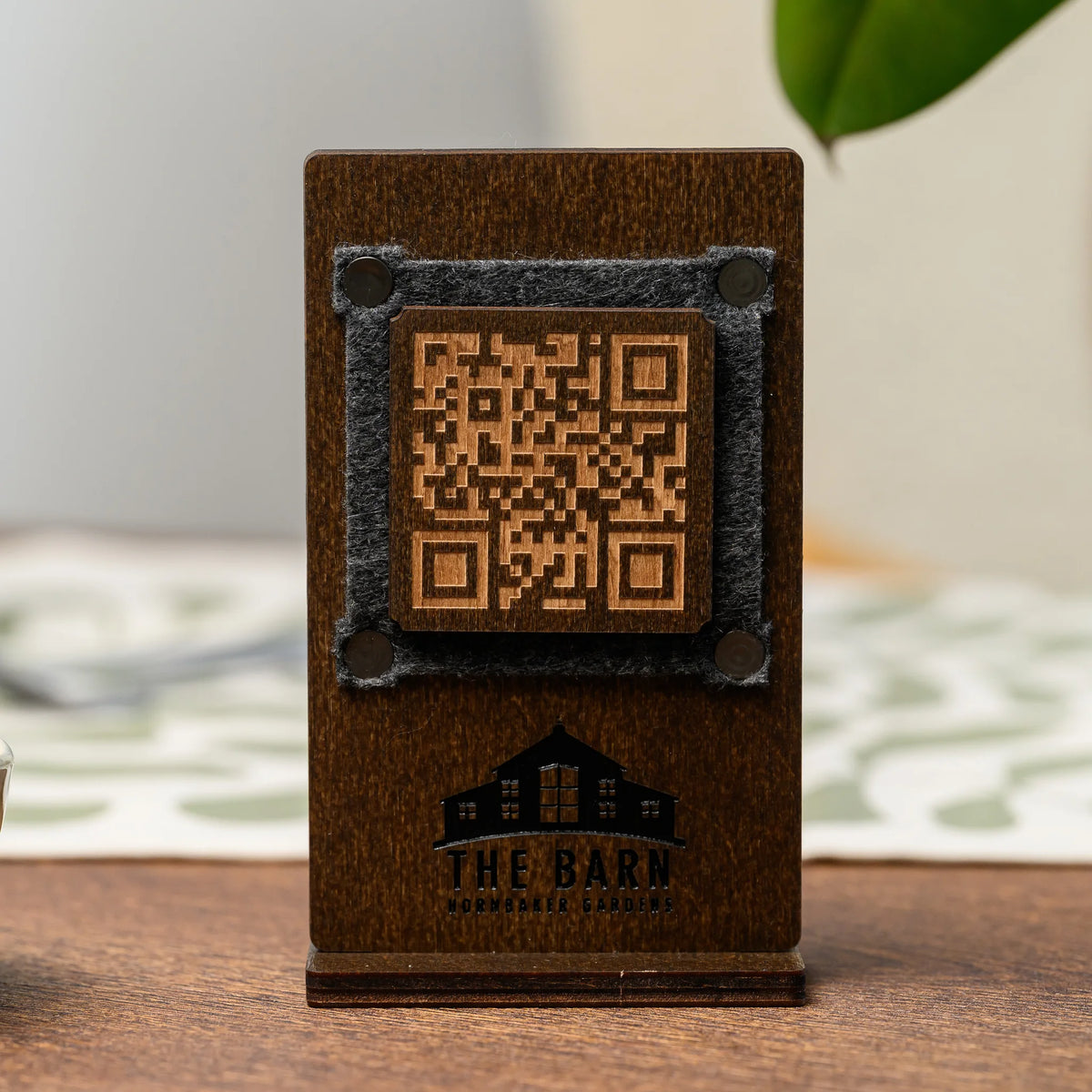 A plywood QR code table stand with a felt frame and an engraved logo, displayed on a bare wooden tabletop
