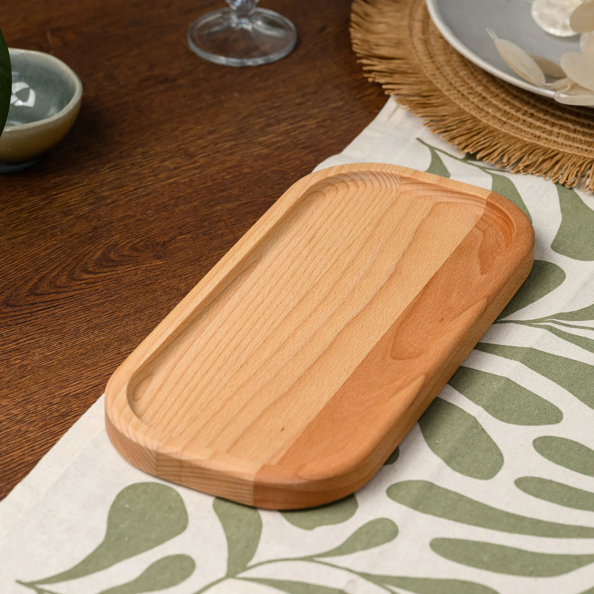 Angled view of an empty beech wood rectangular serving board resting on a leaf-patterned linen tablecloth
