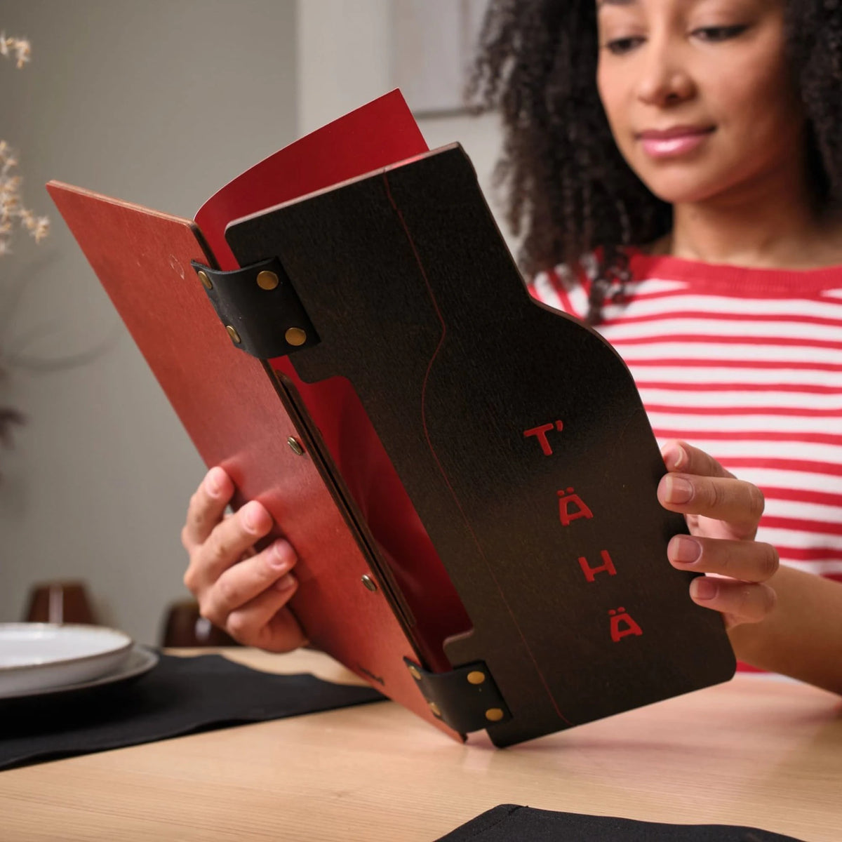 Woman reading open dark wenge plywood slim menu folder with red interior panel and engraved T'AHA logo inside
