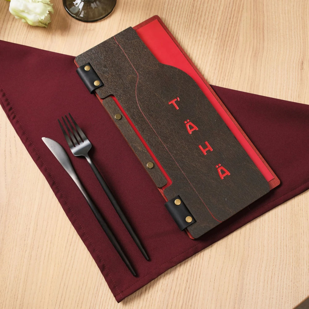 Closed dark wenge plywood slim menu folder with red engraved T'AHA logo, red napkin and black cutlery beside
