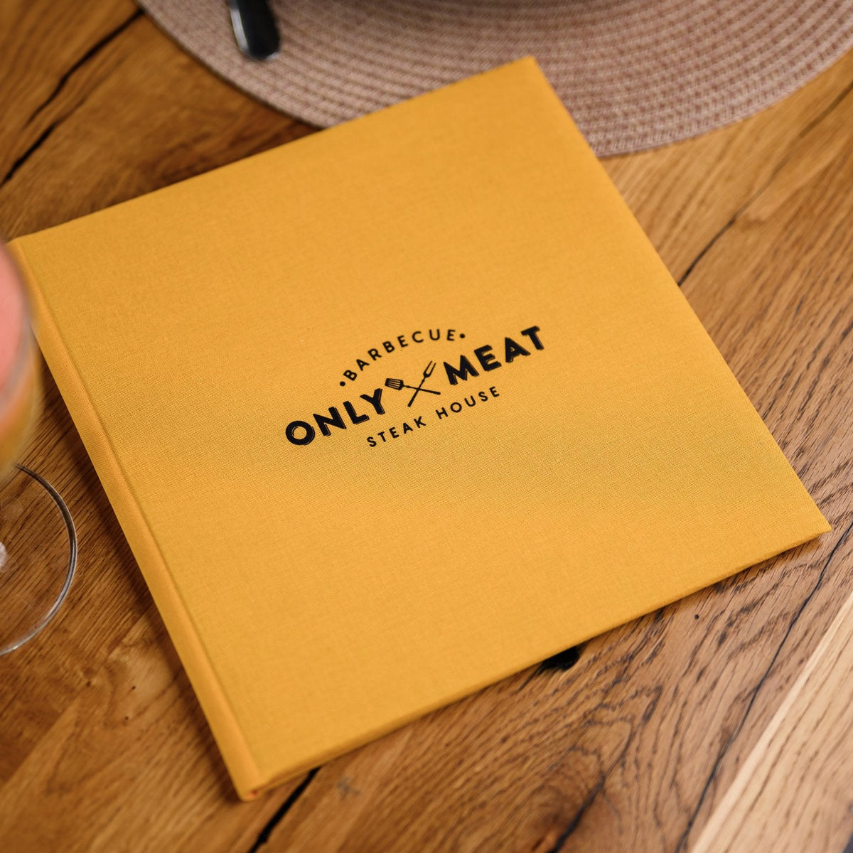 Closed mustard-yellow textile menu folder standing upright with a black logo on a woven table placemat