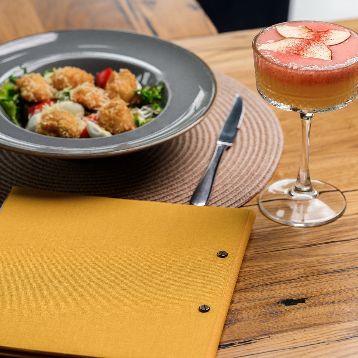 Closed mustard-yellow textile menu folder standing upright beside a salad plate and cocktail on a woven mat