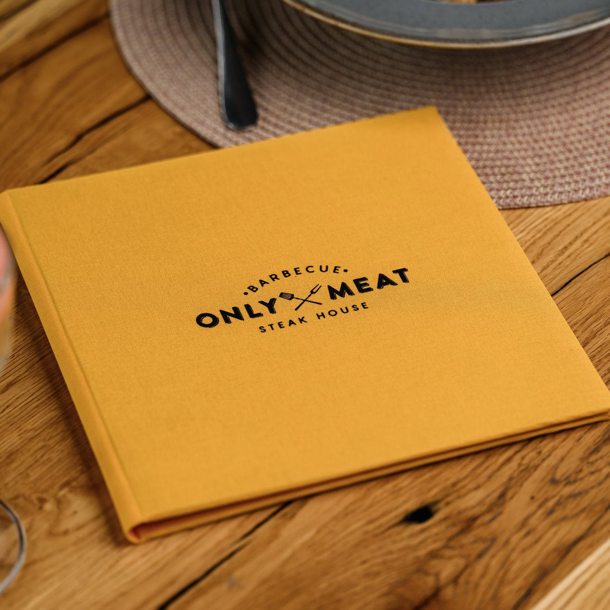 Closed mustard-yellow textile menu folder standing upright with a black logo beside a blurred wine glass