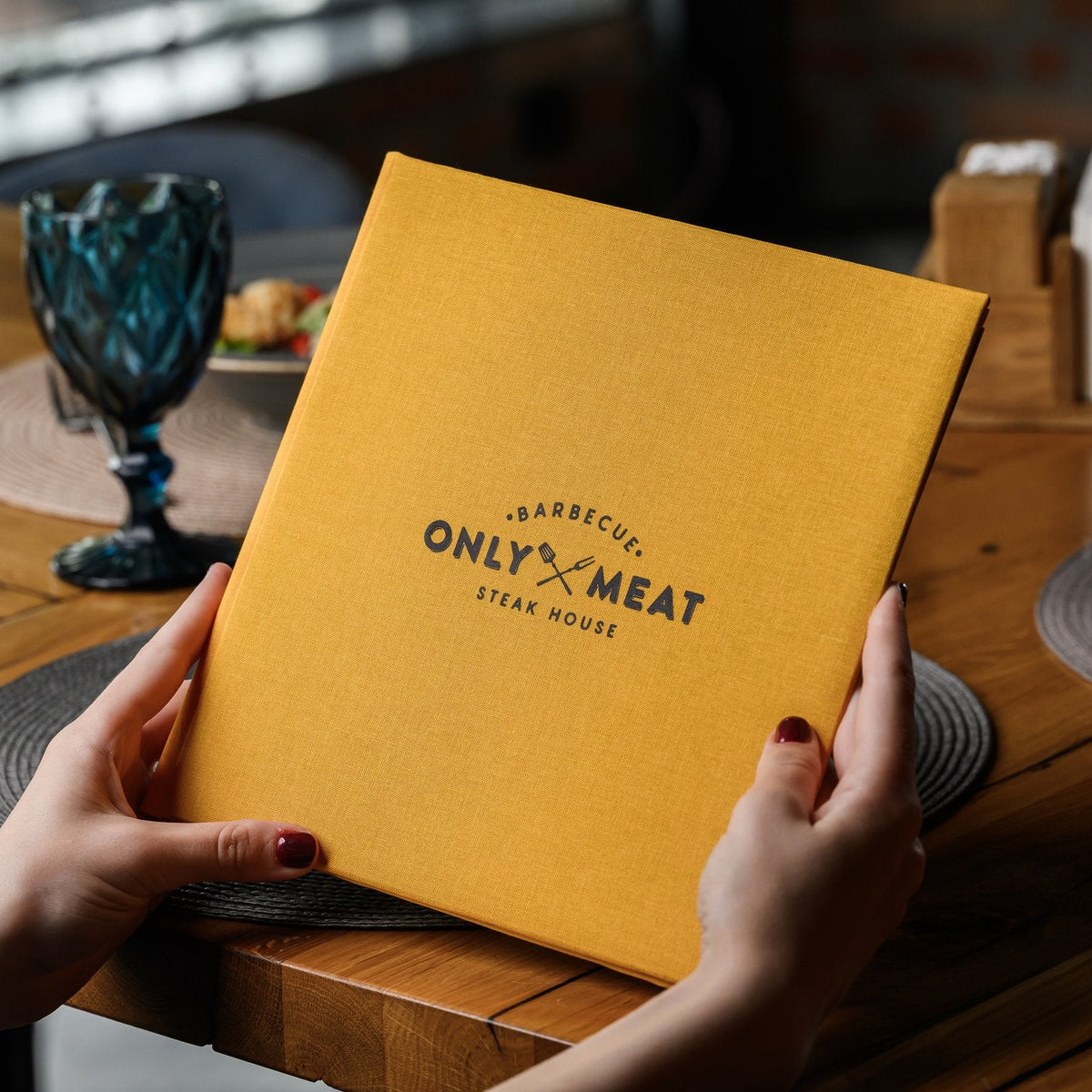 Hand holds a closed mustard-yellow textile menu folder with a black logo standing on a wooden restaurant table