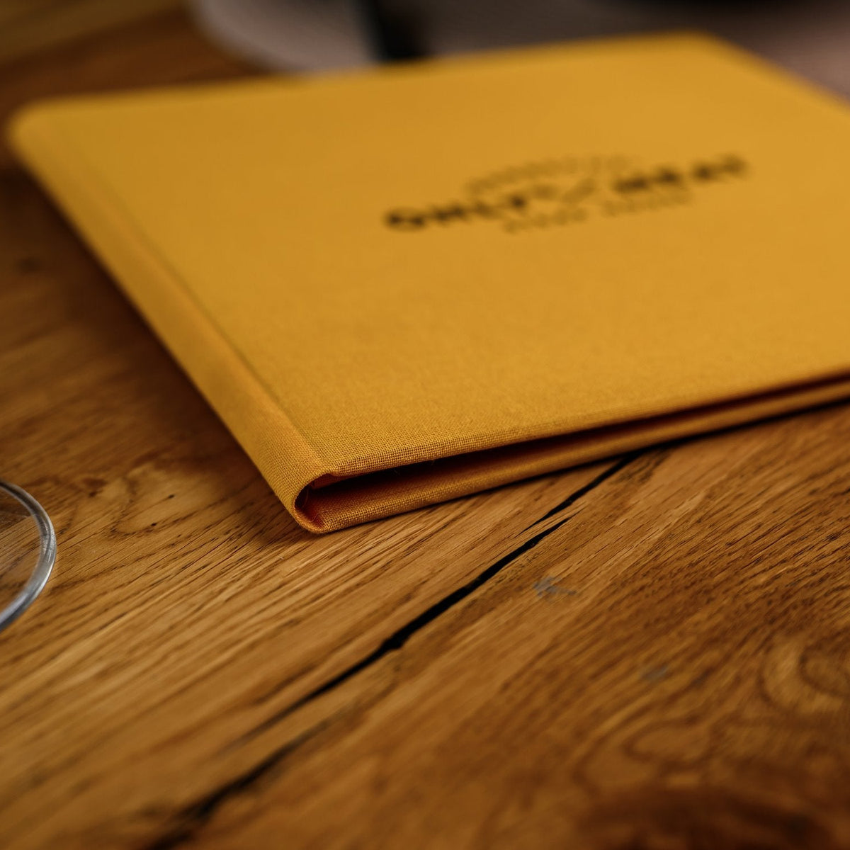 Closed mustard-yellow textile menu folder lying flat on a wooden table with a black printed logo on cover