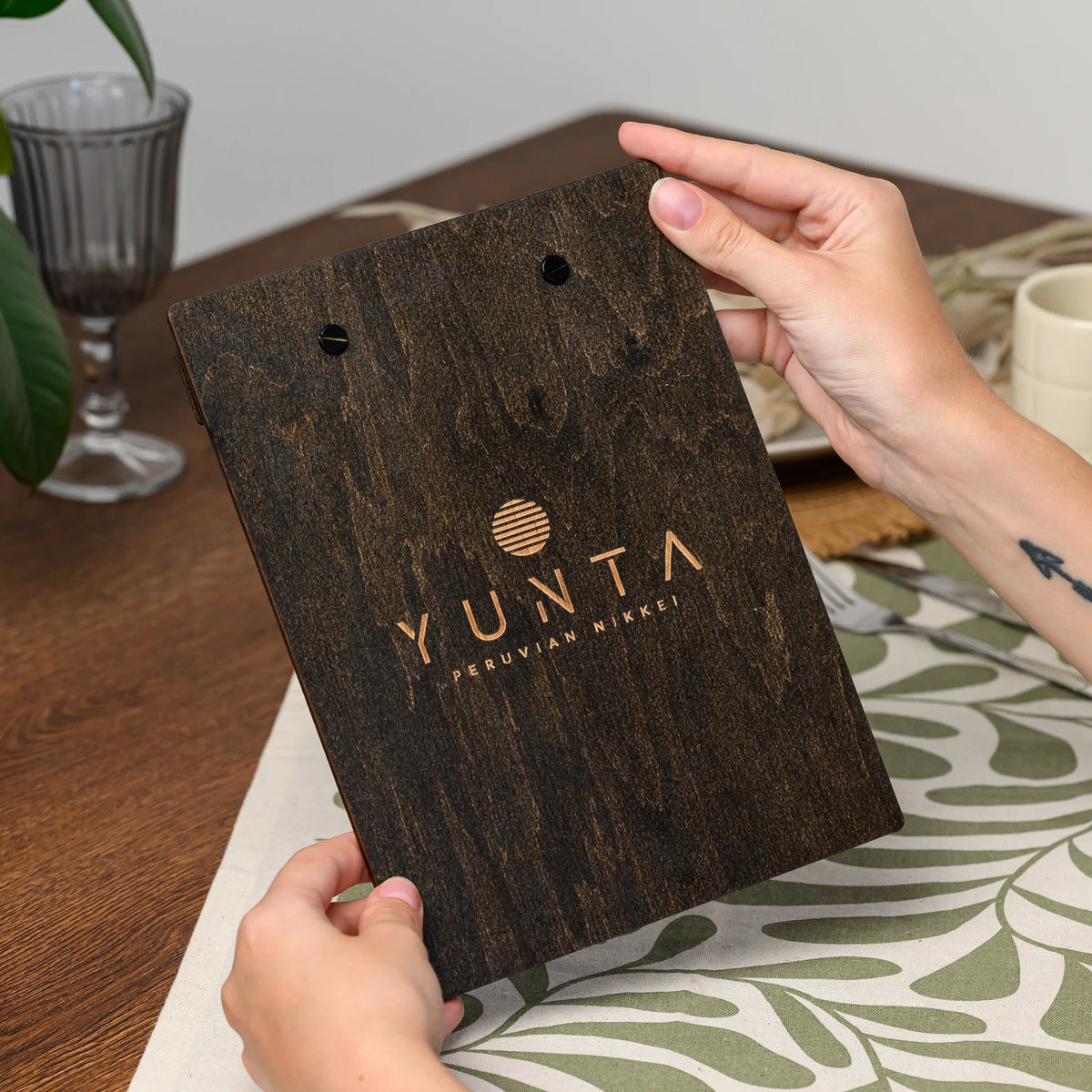 Hands holding the reverse side of a menu board at an angle, showing an engraved logo on the plywood back 